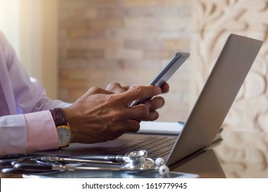 Male doctor in white lab coat hand holding and using mobile smart phone with stethoscope and laptop computer on the desk in medical room at clinic or hospital. Medic tech, online medical concept.