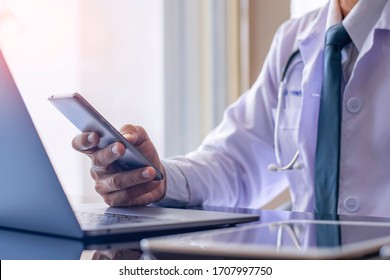 Male doctor in white lab coat, hand holding and using mobile smart phone with laptop computer and digital tablet on the desk. Medical online networking, telehealth, telemedicine, ehr, emr concept.