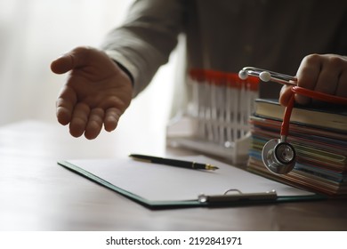 Male Doctor In White Lab Coat Reading Text Book, Search Information And Working On Laptop Computer With  Medical Stethoscope On The Desk In Room At Clinic. Medical Knowledge And Education Concept.