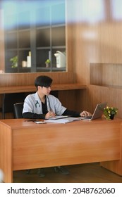 Male Doctor Working At His Office Desk, Writing A Medical Email On Laptop Computer, Checking An Online Prescription Form