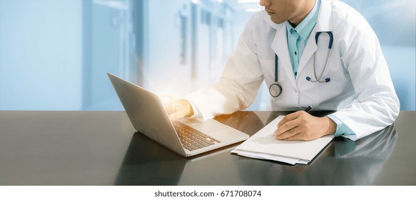 Male Doctor Working On Desk With Laptop Computer And Writing On Paperwork In Office For Medical Data Record.