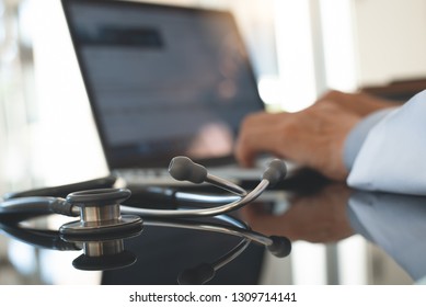 Male doctor working on laptop computer, browsing internet or teleconference with stethoscope on desk in doctor office, close up. Medical and technology, online medical concept, close up