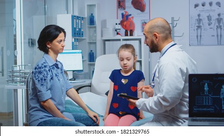 Male Doctor Writing Child Diagnostic On Tablet Talking With Woman. Healthcare Practitioner Physician Specialist In Medicine Providing Health Care Services Consultation Treatment In Hospital