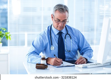 Male Doctor Writing Prescription White Sitting At Desk In Hospital