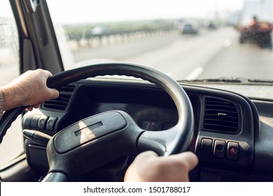 Male Driver Hands Are Holding Steering Wheel Of Truck During The Movement In The Road. Image With Selective Focus On The Wheel, Dashboard And Blurred Windshield