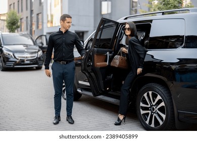 Male driver helps a business lady to get out of a car, opening door of a luxury SUV taxi. Business lady with handbag wearing black formal wear. Concept of transportation service