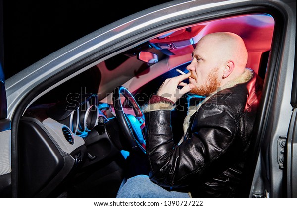 Male driver in a leather jacket\
smoking in the car in the dark. Night unusual photo\
shoot