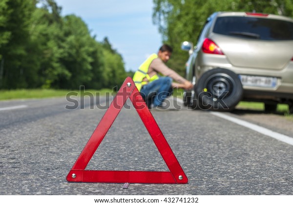 Male driver in reflective vest\
changing tire after breakdown. Focus on red warning triangle\
sign.