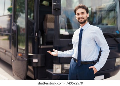 A male driver smiling and posing against a black tourist bus. Behind the back is a modern black tourist bus. On the man the driver's uniform.