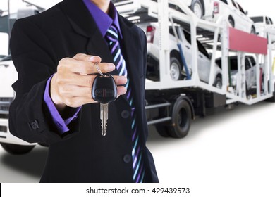 Male driver wearing formal suit and gives a car key with the trailer truck background carrying new cars