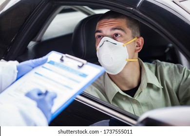 Male driver wearing protective N95 face mask sitting by left drive wheel in UK drive-thru COVID-19 test centre,answering health check up questions,medical worker ticking off symptoms on clipboard form