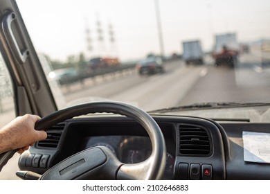 Male Drives Hands Are Holding Steering Wheel Of Truck During The Movement In The Road. Image With Selective Focus On The Wheel, Dashboard And Blurred Windshield