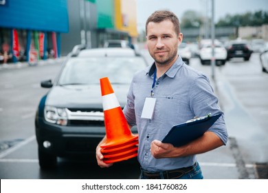 Male driving instructor standing with orange traffic cones and clipboard in his hands. Caucasian man working in driving school. Blurred gray car in background.