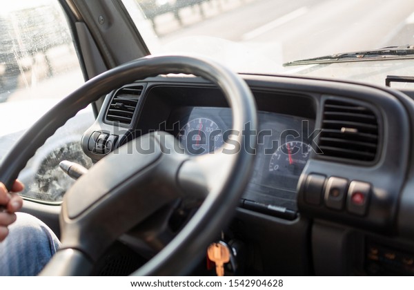 Male driving a truck and holding steering\
wheel of truck during the movement in the road. Image with\
selective focus on the wheel, dashboard with speedometer and\
tachometer, blurred\
windshield