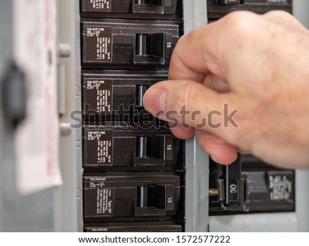 Male electrician turning off power for electrical outlet at circuit breaker box. Resetting tripped breaker in residential electricity power panel.