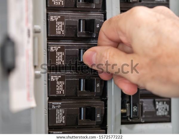 Male electrician turning off power for\
electrical outlet at circuit breaker box. Resetting tripped breaker\
in residential electricity power\
panel.