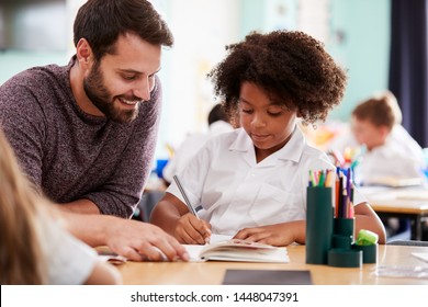 Male Elementary School Teacher Giving Female Pupil Wearing Uniform One To One Support In Classroom