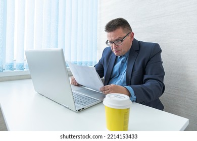 A Male Employee In A Blue Jacket Works With Documents And On A Computer In A Bright Office. Surprised Emotion On The Face.