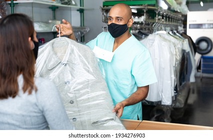 Male Employee In Protective Mask With Client In Modern Laundry, Returning Clothing After Dry Cleaning