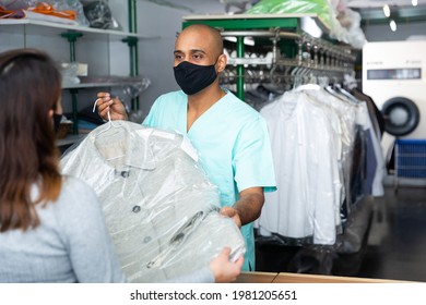 Male Employee In Protective Mask With Client In Modern Laundry, Returning Clothing After Dry Cleaning