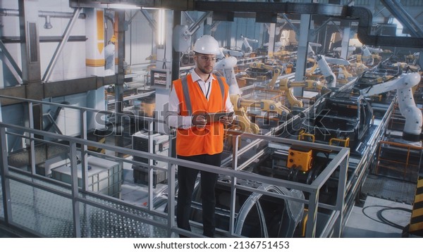 Male employee in\
uniform and hardhat browsing tablet against automatic machines on\
modern vehicle factory