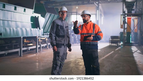 Male employee in uniform and hardhat looking around and showing factory workshop to inspector with tablet