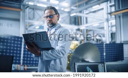 Male Engineer Confident and Focused Thinking, working on Laptop at Aerospace Satellite Manufacturing Facility. Top World Scientists Doing Science and Technology Research in Space Program