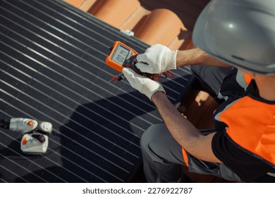 Male Engineer in safety helmet and orange vest testing solar panels voltage with digital multimeter on a city rooftop. Renewable energy concept. Horizontal, close up, top view, copy space - Powered by Shutterstock