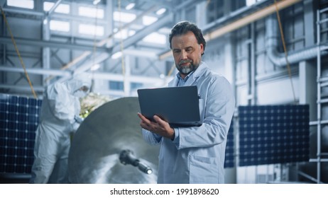 Male Engineer Uses Laptop Computer While Working On Satellite Construction. Aerospace Agency Manufacturing Facility: Scientists Develop, Assemble Spacecraft For Space Exploration, Observation Mission