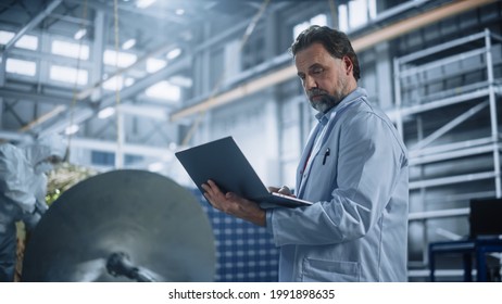 Male Engineer Uses Laptop Computer While Working On Satellite Construction. Aerospace Agency Manufacturing Facility: Scientists Develop, Assemble Spacecraft For Space Exploration, Observation Mission