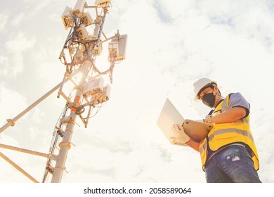 Male engineer wearing a mask working outdoors uses a laptop for maintenance to control the installation and inspection of the telecom tower network communication technology network.