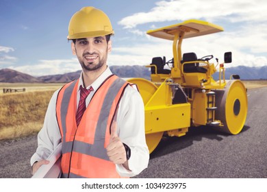 Male Engineer Wearing Safety Helmet Standing In Front Of Steamroller Parked On Road Construction Site