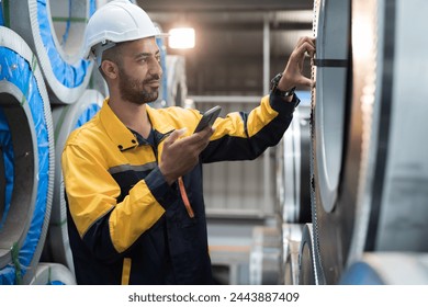 Male engineer worker using digital tablet inspecting quality rolls of galvanized or metal sheet in in aluminum material warehouse. Male worker working in warehouse during distribution process