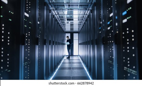 Male IT Engineer Works On A Laptop In A Big Data Center. Rows Of Rack Servers Are Seen.