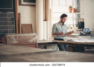 Male entrepreneur craftsman checking orders and figures on a clipboard while standing casually in his studio workshop