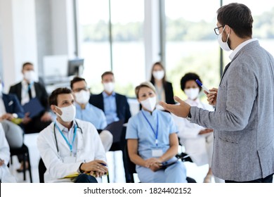 Male entrepreneur holding a training class to large group of healthcare workers and business people in conference hall. They are all wearing protective face mask due to COVID-19 pandemic. 