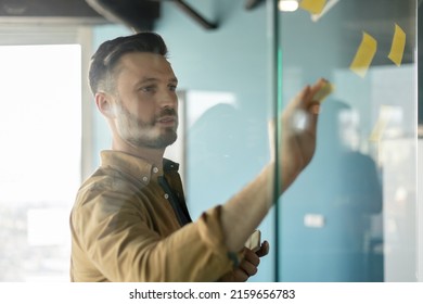 Male Entrepreneur Putting Sticky Notes On Glass Wall Standing In Modern Office. Businessman Planning Project Working At Workplace. Successful Management And Entrepreneurship. Selective Focus