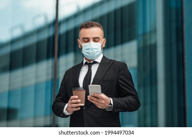 Male Entrepreneur In Suit Wearing Face Mask And Using Smartphone, Going To Office And Drinking Morning Coffee, Standing Outdoors Near Glass Building. Back To Office Work After Covid-19 Pandemic