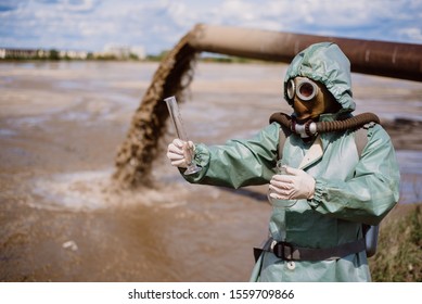 A Male Environmentalist In A Protective Suit And Gas Mask Takes A Sample Of Water. Scientist Doing Toxicological Study.