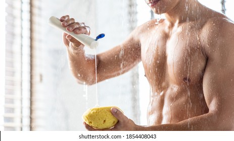 Male Everyday Hygiene And Bodycare Concept. Unrecognizable Man Pouring Shower Gel On Sponge Washing Body Standing Under Falling Hot Water Drops In Modern Bathroom At Home. Banner, Copy Space
