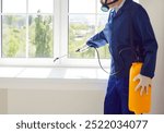 Male exterminator from pest control service wearing protective safety mask and blue workwear suit working inside house spraying toxic liquid or gas from yellow sprayer bottle over white window sill