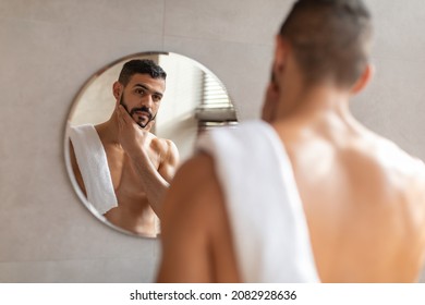 Male Facial Care. Portrait Of Handsome Arab Man Looking At His Reflection In Mirror In Bathroom And Touching Beard, Attractive Masculine Middle Eastern Guy Enjoying His Appearance, Selective Focus