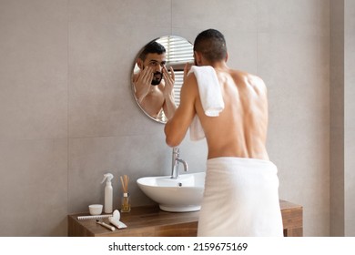 Male Facial Care. Portrait Of Handsome Arab Man Looking At His Reflection In Mirror In Bathroom Touching Undereye Area, Attractive Masculine Middle Eastern Guy Applying Cream, Over The Shoulder View