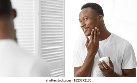 Male Facial Skincare. Black Man Using Moisturizing Aftershave Cream Caring For Face Skin Standing Near Mirror In Bathroom At Home. Men's Beauty And Self-Care Routine Concept. Panorama