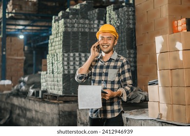 male factory worker smiles while making a phone call and carrying a clipboard in a storage warehouse - Powered by Shutterstock