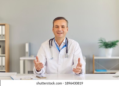 Male Family Doctor Sits In Front Of The Camera And Gives An Online Consultation To His Patient While Sitting In The Office. Friendly Doctor In A Medical Gown Explains The Patient's Diagnosis.