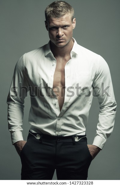 Male fashion,\
beauty concept. Portrait of brutal young man with short wet blond\
hair wearing white shirt, black pants, posing over gray background.\
Classic style. Studio shot