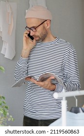 A Male Fashion Designer Or Fashion Designer Flips Through A Magazine And Talks On The Phone With A Client. An Adult Business Man Is Working In His Office. Senior Creative Director In A Striped Jacket