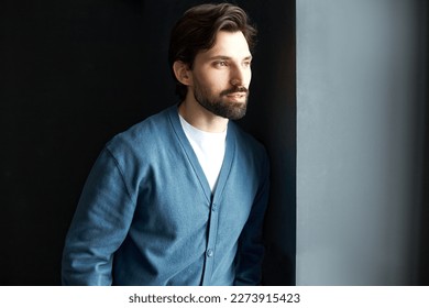 Male fashion and menswear. Side view of pensive romantic-looking guy of 30 with beard on chin wearing blue classical cardigan standing against black studio background leaning on gray wall