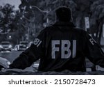 Male FBI agent wearing dark blue coat with FBI logo looking down the street with cars in the dusk seen from behind.